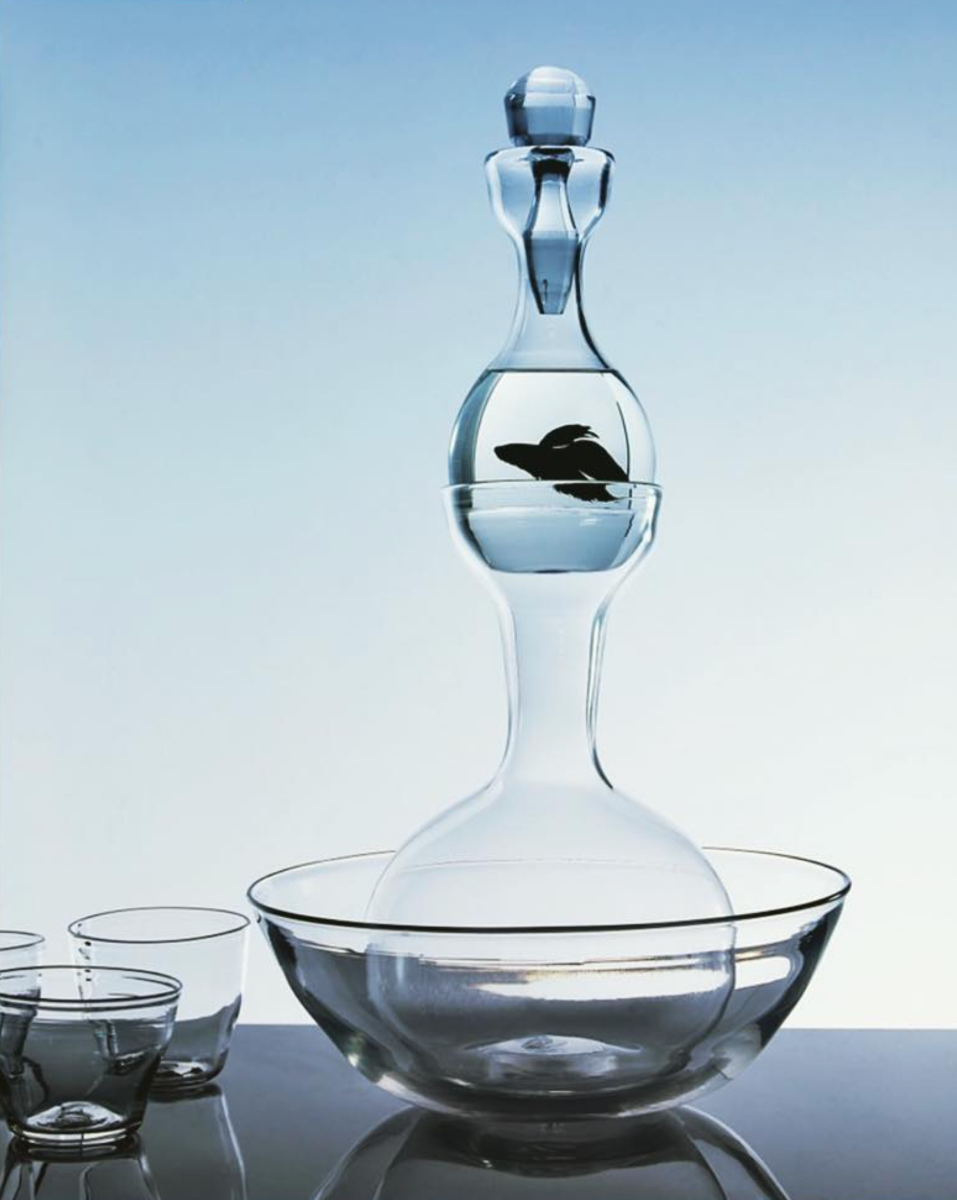 Clear glass decanter with a bowl at the bottom on a reflective surface with a light blue gradient background.